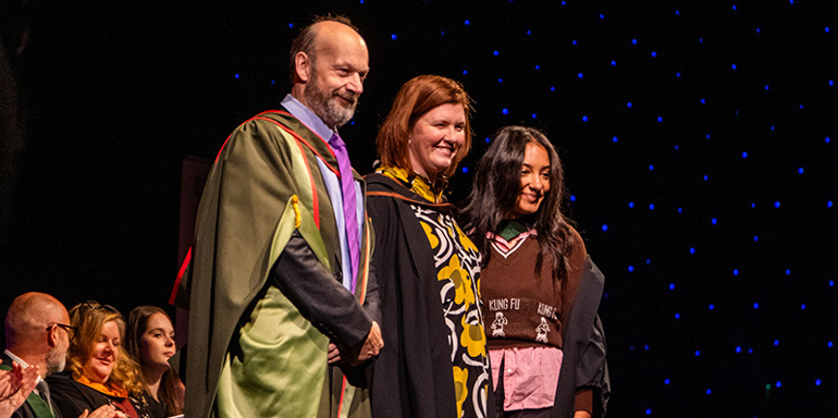 Professor John Craig (Pro Vice Chancellor, University of Hull), Patsy Gilbert (Vice Principal, Leeds Conservatoire) and Lily Fontaine (frontwoman of English Teacher) on stage at Graduation 2025 in New Dock Hall, Royal Armouries