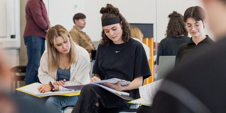 Students in a classroom at their desks reading a script