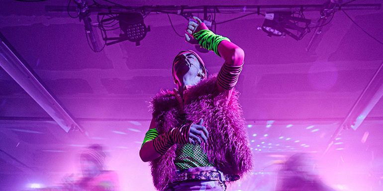 Students on stage wearing colourful attire with purple lighting. In School of Drama Production of A Midsummer Night's Dream